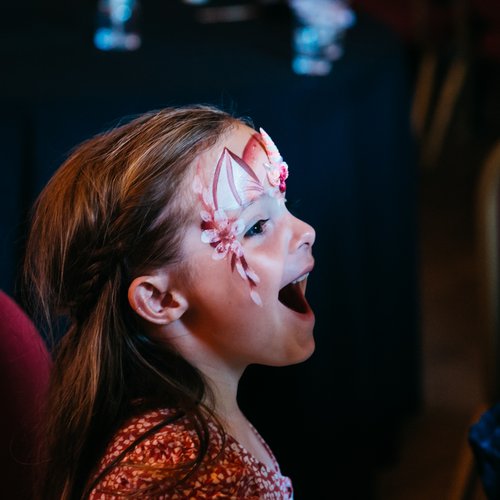 A child smiling at seeing her face painted in the mirror at the Solving Kids' Cancer UK Family Fun Day.
